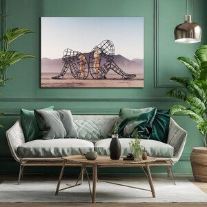 May include: A large canvas art print depicting two wireframe figures embracing on a desert landscape. The artwork is displayed above a modern sofa with green and gray throw pillows, enhancing the room's aesthetic. The room is painted in a teal green color.