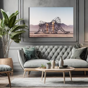 May include: A framed canvas art piece depicting two wireframe figures in a desert landscape. The artwork hangs above a gray tufted sofa with wooden legs, complemented by a round coffee table and a large potted plant.