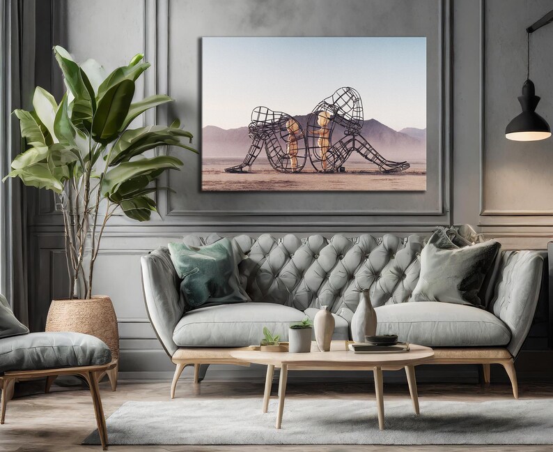 May include: A framed canvas art piece depicting two wireframe figures in a desert landscape. The artwork hangs above a gray tufted sofa with wooden legs, complemented by a round coffee table and a large potted plant.