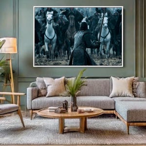 May include: A living room scene featuring a gray sectional sofa, wooden armchair, and a round coffee table on a patterned rug. A large framed artwork depicting a battle scene hangs above the sofa. A floor lamp and a side table with a lamp provide lighting.