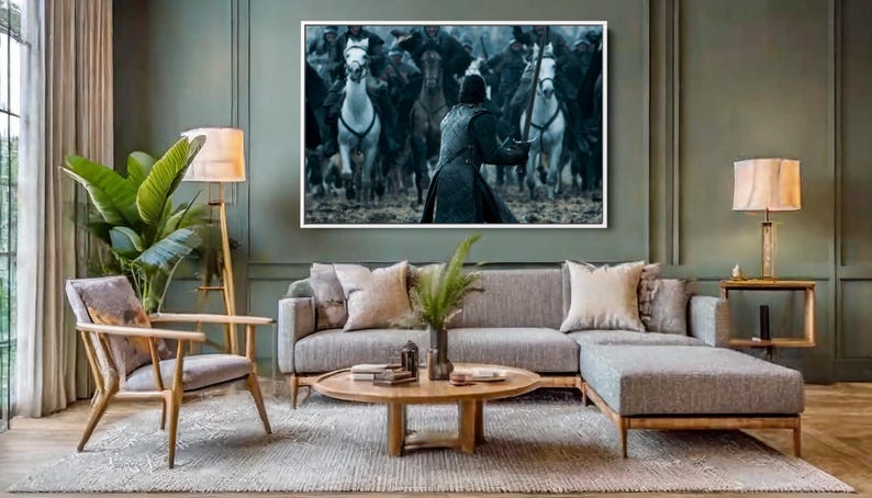 May include: A living room scene featuring a gray sectional sofa, wooden armchair, and a round coffee table on a patterned rug. A large framed artwork depicting a battle scene hangs above the sofa. A floor lamp and a side table with a lamp provide lighting.