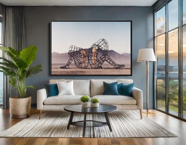 May include: A living room scene featuring a large framed artwork depicting two wireframe figures in a seated position. The room includes a beige sofa with blue and white pillows, a round black coffee table, and a floor lamp.
