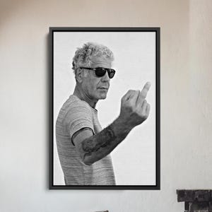 May include: Black and white framed print featuring a man wearing sunglasses and a striped t-shirt, extending his middle finger. The image is a portrait, with the subject's arm and a tattoo visible. The background is plain white.