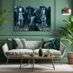 May include: A framed canvas print depicting a battle scene with riders on horseback. The artwork features a central figure facing a charge of horses, with a muted color palette of blues, grays, and whites. The print is displayed above a modern sofa.