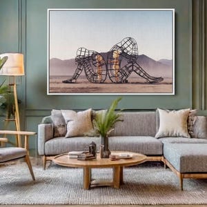 May include: A living room scene featuring a large framed artwork depicting two wireframe figures in a desert landscape. The room includes a gray sectional sofa, wooden chair, and a round coffee table. A floor lamp and a table lamp provide lighting.