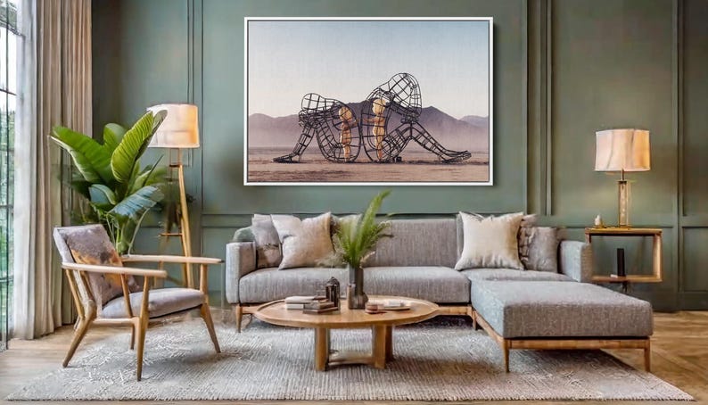 May include: A living room scene featuring a large framed artwork depicting two wireframe figures in a desert landscape. The room includes a gray sectional sofa, wooden chair, and a round coffee table. A floor lamp and a table lamp provide lighting.