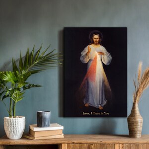 May include: A framed canvas print of a religious figure with a halo, wearing a white robe, and radiating light. The text "Jesus, I Trust in You" is at the bottom. The artwork is displayed on a wooden table with a plant and decorative vase.