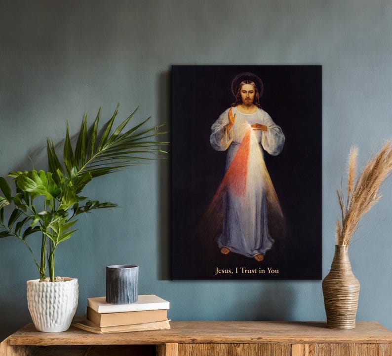 May include: A framed canvas print of a religious figure with a halo, wearing a white robe, and radiating light. The text "Jesus, I Trust in You" is at the bottom. The artwork is displayed on a wooden table with a plant and decorative vase.