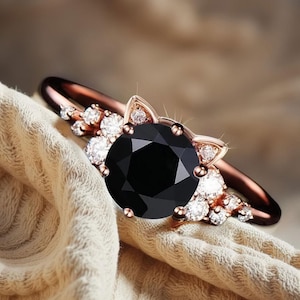 Könnte beinhalten: Ein roségoldener Ring mit einem großen, facettierten schwarzen Edelstein im Mittelpunkt. Der Ring hat ein Katzenohren-Design mit kleinen, klaren Edelsteinen und zusätzlichen klaren Edelsteinen am Band. Der Ring ist vor einem neutralfarbenen Stoff platziert.