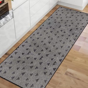 Könnte beinhalten: Ein rechteckiger Küchenläufer mit einem geometrischen Muster aus schwarzen Dreiecken und Linien auf grauem Hintergrund. Der Teppich liegt auf einem Hartholzboden, und die Ränder sind schwarz. Der Teppich befindet sich in der Nähe eines weißen Küchenschranks.