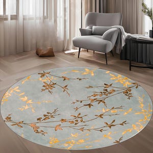 May include: A round area rug with a light blue background and a floral design in shades of gold and brown. The rug is placed on a wooden floor, with a gray armchair and side table in the background.