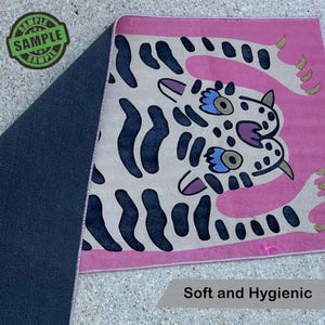 Puede incluir: Una alfombra rosa con un dise&ntilde;o de tigre de dibujos animados. El tigre tiene manchas negras, ojos azules y morados y patas color canela. La alfombra tiene un reverso azul oscuro y las palabras "Soft and Hygienic" est&aacute;n impresas.