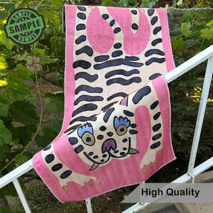 Puede incluir: Alfombra decorativa rosa y crema con un dise&ntilde;o de tigre estilizado. La alfombra tiene un borde rosa y un tigre crema con manchas y rayas negras. El tigre tiene ojos azules y garras rosas. La alfombra est&aacute; colgada en una barandilla blanca.