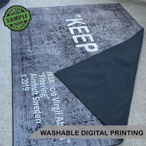 Puede incluir: Una alfombra rectangular gris con un patr&oacute;n floral desgastado y la palabra "KEEP" en letras blancas grandes. La alfombra tiene un reverso gris oscuro e incluye el texto "IKEA c/o Virgil Ab" y "Washable Digital Printing."