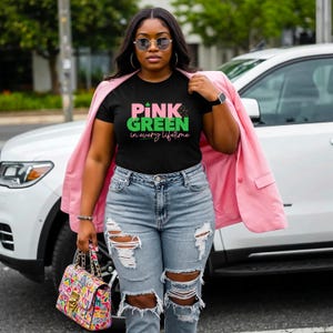Może przedstawiać: Czarny t-shirt z napisem "PINK & GREEN in every lifetime" w kolorze różowym i zielonym. Koszulka jest noszona z jasnoniebieskimi podartymi dżinsami i różową marynarką. W dłoni trzymana jest kolorowa torebka.