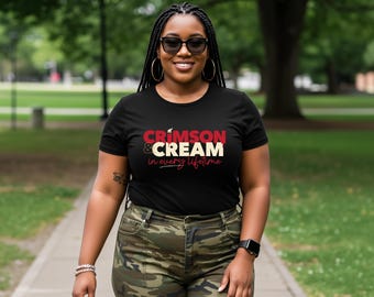 Op de afbeelding: Een zwart t-shirt met de tekst "CRIMSON & CREAM in every lifetime" in rode en crèmekleurige kleuren. De persoon draagt een camouflagebroek en een zonnebril. De achtergrond is een park met bomen.