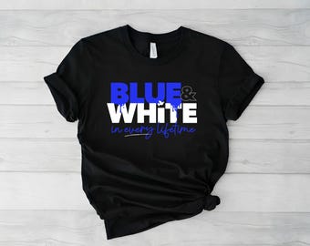 Op de afbeelding: Zwart T-shirt met de tekst "BLUE & WHITE" in blauwe en witte letters. Daaronder staat de zin "in every lifetime" in blauw schrift. Het T-shirt is opgevouwen op een licht houten oppervlak.