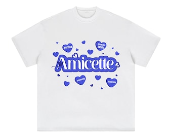 Op de afbeelding: Wit t-shirt met het woord "Amicette" in blauw schrift, omringd door blauwe harten en tekst. Het shirt bevat ook de woorden "service", "scholarship" en "leadership".