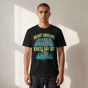 Pode incluir: Camiseta preta com um design gráfico com esqueletos e o texto "HEART SAYS PIT KNEES SAY SIT" em verde e branco. O design inclui chamas azuis ao redor dos esqueletos.