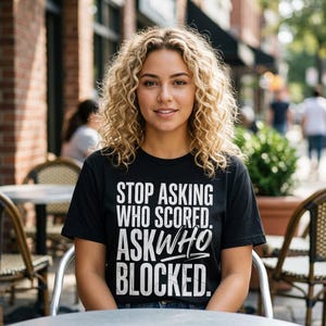 May include: A black short-sleeved t-shirt featuring white text that reads: STOP ASKING WHO SCORED. ASK WHO BLOCKED. The lettering is arranged in a bold, stacked layout with a distressed texture on the front of the garment.