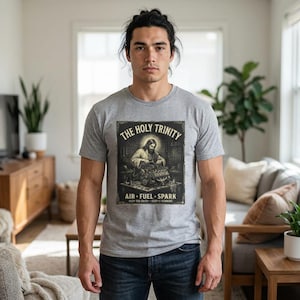 Könnte beinhalten: Ein graues T-Shirt mit einem Aufdruck. Der Aufdruck zeigt den Text "THE HOLY TRINITY" über einem Bild von Jesus, der an einem Motor arbeitet. Unter dem Bild steht der Text "AIR - FUEL - SPARK", mit "KEEP THE FAITH - KEEP IT RUNNING" am unteren Rand.
