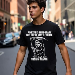 May include: Black t-shirt with the text "PINKEYE IS TEMPORARY BUT SHE'LL NEVER FORGET" and "THE RIM REAPER". The graphic features a Grim Reaper holding a scythe. The person is standing on a city street.