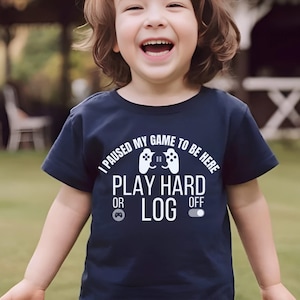 以下が含まれることがあります： 白い文字で「I paused my game to be here. Play hard or log off」と書かれたネイビーブルーのTシャツ。コントローラーのグラフィックが特徴です。子供がそのシャツを着て笑顔です。