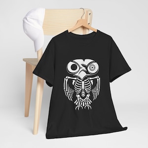 May include: Black t-shirt featuring a white owl skeleton design. The graphic includes the owl's eyes, beak, rib cage, wings, and feet. A white baseball cap hangs on a wooden chair in the background.
