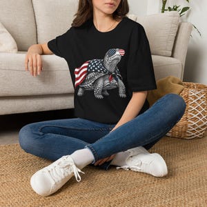 Könnte beinhalten: Schwarzes T-Shirt mit einer detaillierten Illustration einer Schildkröte, die eine Sonnenbrille und einen Umhang mit dem Design der amerikanischen Flagge trägt. Die Schildkrötengrafik ist in Grau- und Weißtönen gehalten, mit roten und blauen Akzenten. Das T-Shirt wird von einer Person getragen, die auf einer gewebten Matte sitzt.