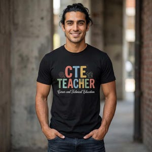 May include: Black t-shirt with the text "CTE TEACHER" in colorful letters. The shirt also includes the words "Career and Technical Education" in a smaller font below the main text. The person is smiling and wearing the shirt.