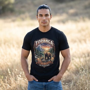 Op de afbeelding: Zwart t-shirt met een afbeelding van een beer in een berglandschap, het woord "TOPANGA" bovenaan en de tekst "COME FOR THE VIBE STAY FOR THE BEARS" eronder. Het t-shirt wordt door een persoon gedragen.