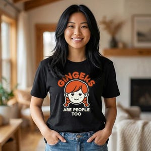 May include: Black t-shirt with the text "Gingers Are People Too" in orange and white. The shirt features a cartoon illustration of a redhead with pigtails. The person in the image is wearing blue jeans.