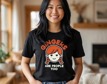 May include: Black t-shirt with the text "Gingers Are People Too" in orange and white. The shirt features a cartoon illustration of a redhead with pigtails. The person in the image is wearing blue jeans.