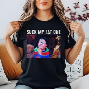 Puede incluir: Camiseta negra con el texto "SUCK MY FAT ONE" sobre una imagen de un niño con gafas y una camiseta a rayas de colores, mostrando el dedo medio.