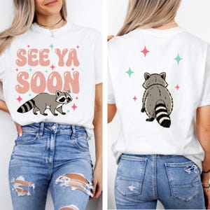 Puede incluir: Camiseta blanca con un diseño de mapache y el texto "SEE YA SOON" en rosa y verde claro. La parte trasera de la camiseta presenta un diseño de mapache y estrellas. La camiseta se combina con vaqueros azules.