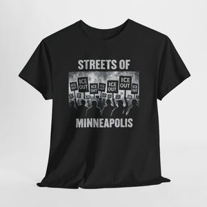 Könnte beinhalten: Schwarzes T-Shirt mit den weißen Worten "STREETS OF MINNEAPOLIS". Das Shirt zeigt eine Grafik von Personen, die Schilder mit der Aufschrift "ICE OUT" halten.