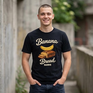 以下が含まれることがあります： バナナとパンのグラフィックと、ヴィンテージスタイルのフォントで「Banana Bread Baddie」という文字が入った黒いTシャツ。柔らかい素材で作られています。