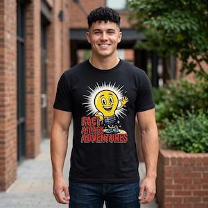 Puede incluir: Una camiseta negra de manga corta con un gráfico de una bombilla amarilla sonriente con brazos y piernas. Debajo de la bombilla, el texto FACT ATTACK ADVENTURES aparece en letras rojas negritas con un borde amarillo sobre un fondo oscuro.