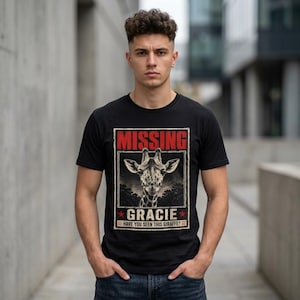 May include: A black t-shirt featuring a graphic print of a giraffe head centered within a rectangular frame. The text reads MISSING at the top, GRACIE in the middle, and HAVE YOU SEEN THIS GIRAFFE? at the bottom, accented by two red stars.