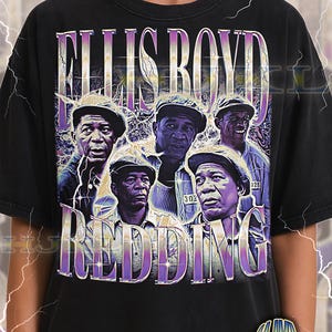 May include: Black adult unisex t-shirt featuring a graphic of five men with the text "ELLIS BOYD REDDING" in a purple and white vintage style. The shirt has a distressed look.