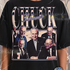 May include: Black adult unisex t-shirt featuring a graphic of Chuck Schumer with the word "CHUCK" in large, chrome-style letters above his name. The shirt has a vintage, distressed look.
