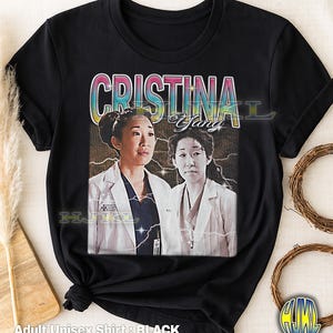 May include: Black adult unisex t-shirt featuring a vintage-style image of Cristina Yang from Grey's Anatomy. The design includes a photo of the character and the name "Cristina Yang" in a colorful, retro font. The shirt is made of a soft material.