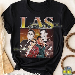 Op de afbeelding: Zwart unisex T-shirt voor volwassenen met een vintage-stijl afbeelding van vier mannen met microfoons. De afbeelding bevat het woord "LAS" in goud en geel, met het woord "Galacticos" eronder. Het shirt heeft een ronde hals en korte mouwen.