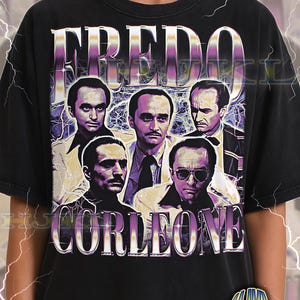 May include: Black adult unisex t-shirt featuring a graphic design with the word "FREDO" above a group of men's portraits and the word "CORLEONE" below. The shirt has a vintage style.