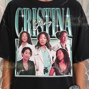 May include: Black adult unisex t-shirt featuring a graphic of the character Cristina Yang. The design includes the name "Cristina Yang" in a teal font, with multiple images of the character. The shirt also has the text "ADULT Unisex Shirt : BLACK".