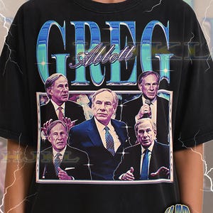 Puede incluir: Camiseta unisex negra para adultos con un diseño gráfico. El diseño presenta el nombre "GREG" en letras grandes, azules y estilizadas sobre una imagen enmarcada de un hombre con traje. La camiseta también incluye el nombre "Abbott" en una fuente cursiva.