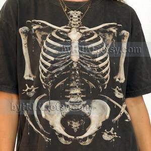 May include: Dark gray t-shirt featuring a detailed, distressed graphic of a human skeleton. The skeleton design is in shades of white and gray, with visible rib cage, spine, and pelvis. The text "Comfort Colors : GRAPHITE" is at the bottom.