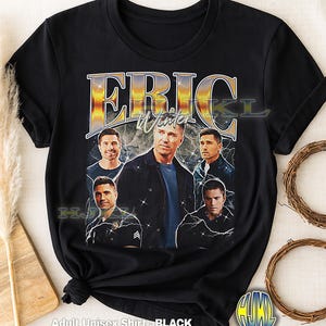 May include: Black adult unisex t-shirt featuring a vintage-style graphic of Eric Winter with a sunset-themed background and lightning bolts. The text "ERIC Winter" is prominently displayed. The shirt is made of a soft material.