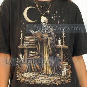 May include: Dark gray t-shirt featuring a skeleton reading a book in a mystical scene. The design includes a crescent moon, stars, candles, and books. The text "Comfort Colors: GRAPHITE" is at the bottom.