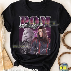 Könnte beinhalten: Schwarzes Unisex-T-Shirt für Erwachsene mit einem Retro-Grafikdesign von zwei Frauen, dem Text "POM" und "Klementieff". Das Design verwendet Lila und Weiß mit Blitz-Akzenten. Das T-Shirt ist aus weichem Material. Der Text "Adult Unisex Shirt: BLACK" steht unten.